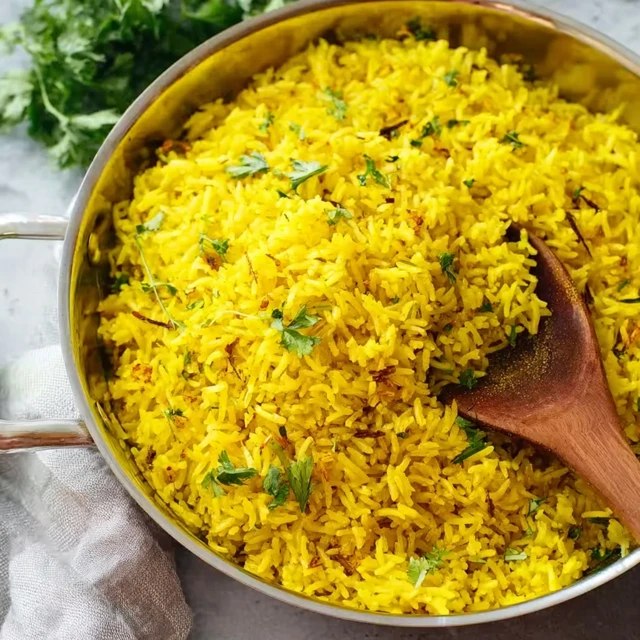 Close-up of fluffy turmeric basmati rice showing each long grain stained bright yellow, fresh green herb confetti, golden brown toasted nuts, glistening with olive oil, lemon wedge beside